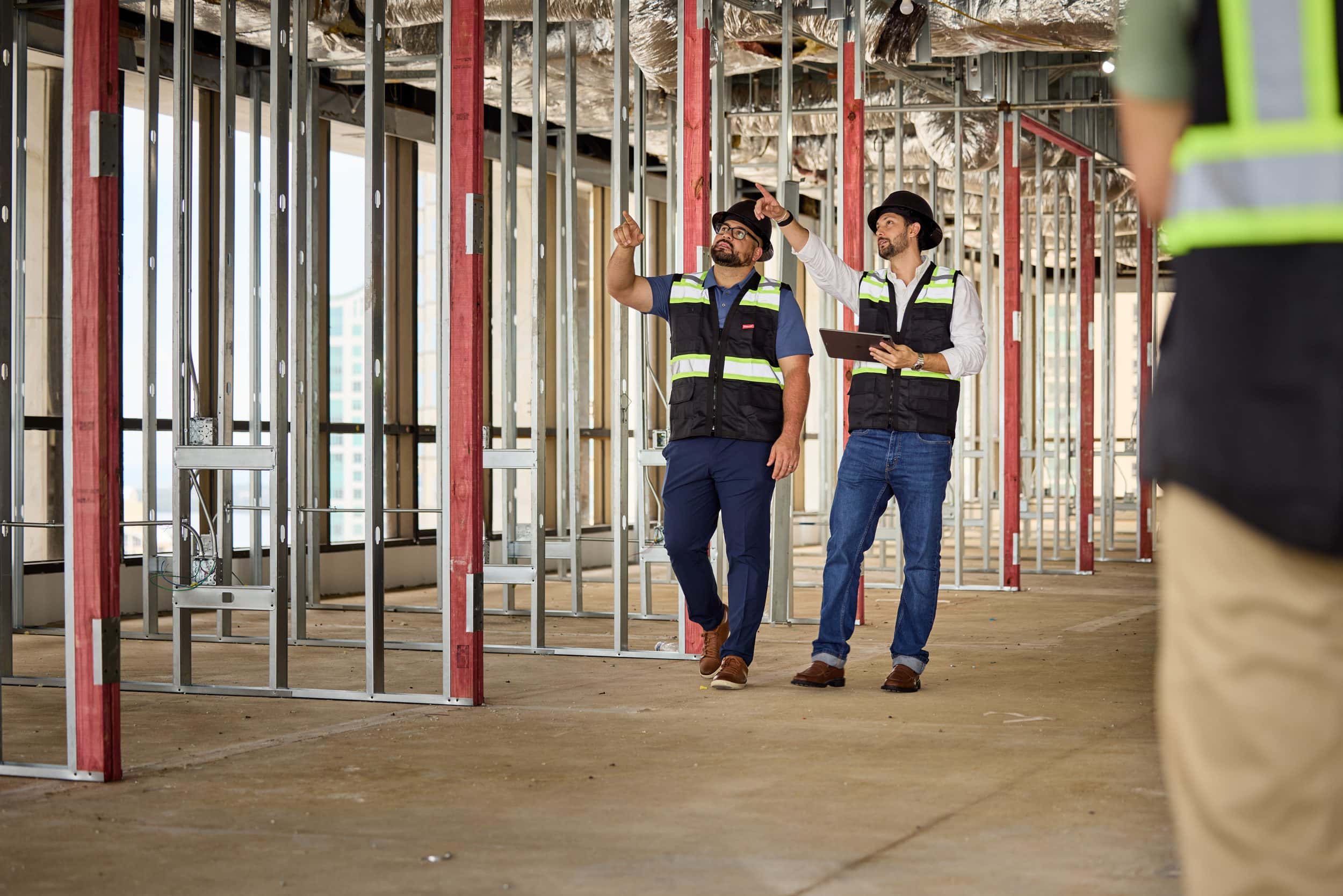 Two SPARC project managers in hard hats pointing up at the ceiling of a floor under construction, metal stud framing on both sides
