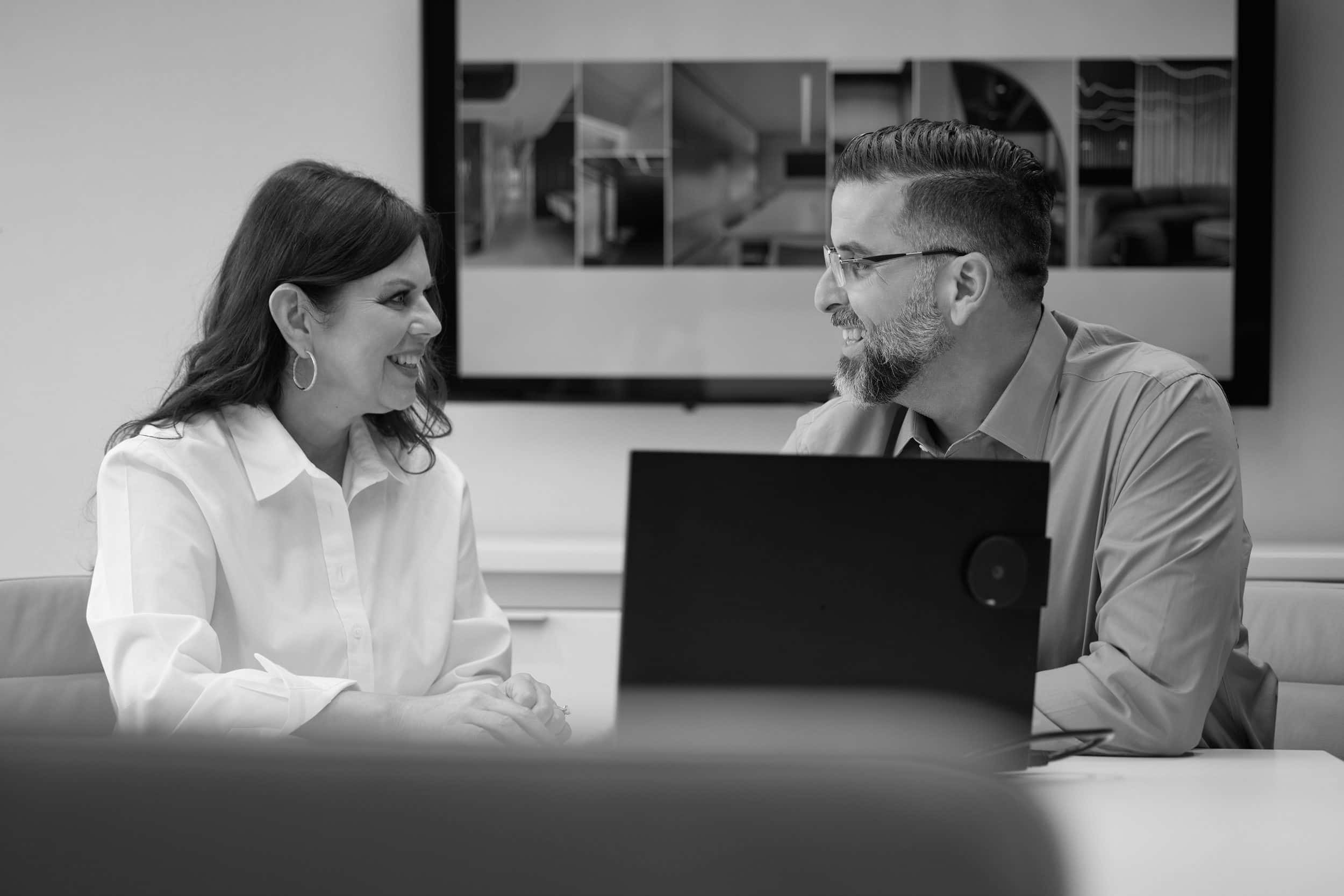 Two members of SPARC leadership in conversation across a conference table, a laptop open between them and project renderings on the wall screen, in black and white
