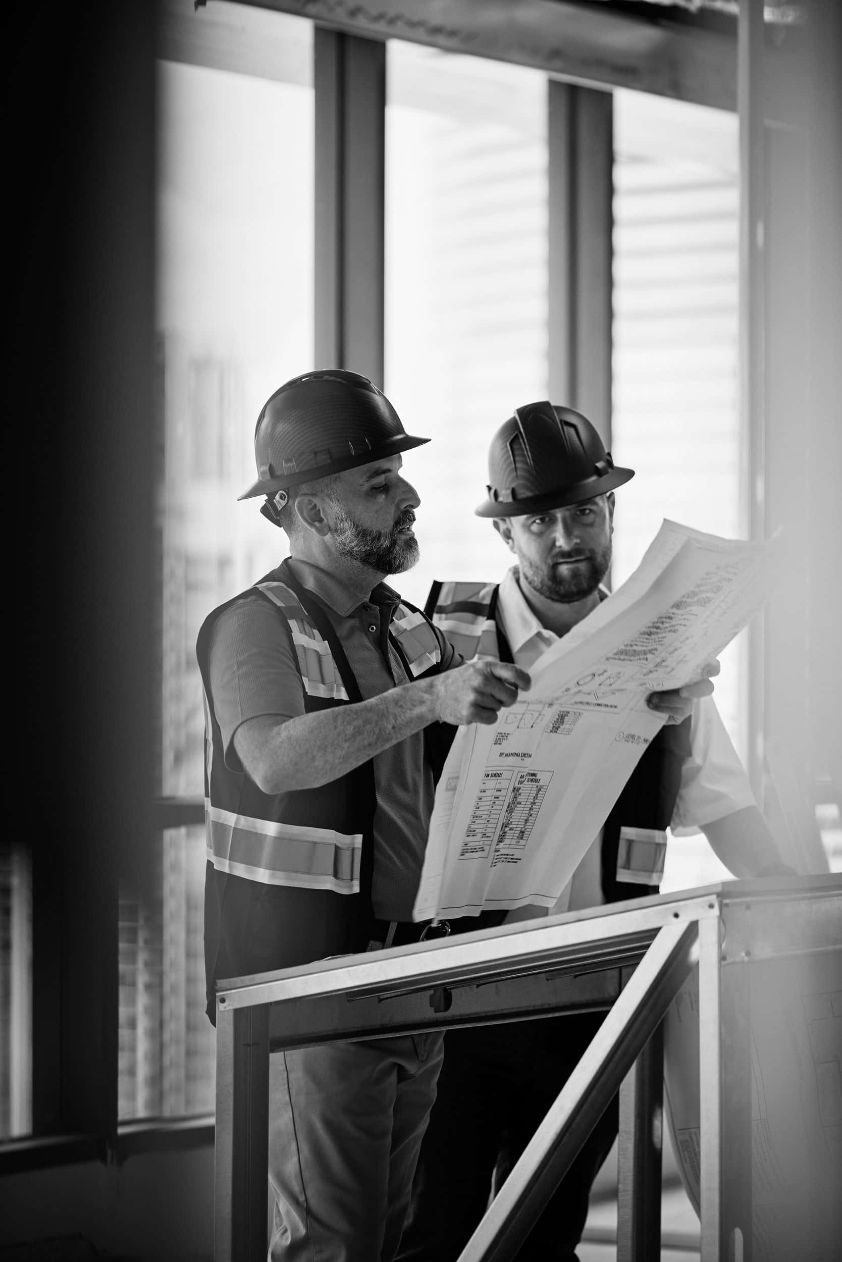 Two SPARC project managers in hard hats study a set of drawings at a window on a floor under construction, in black and white