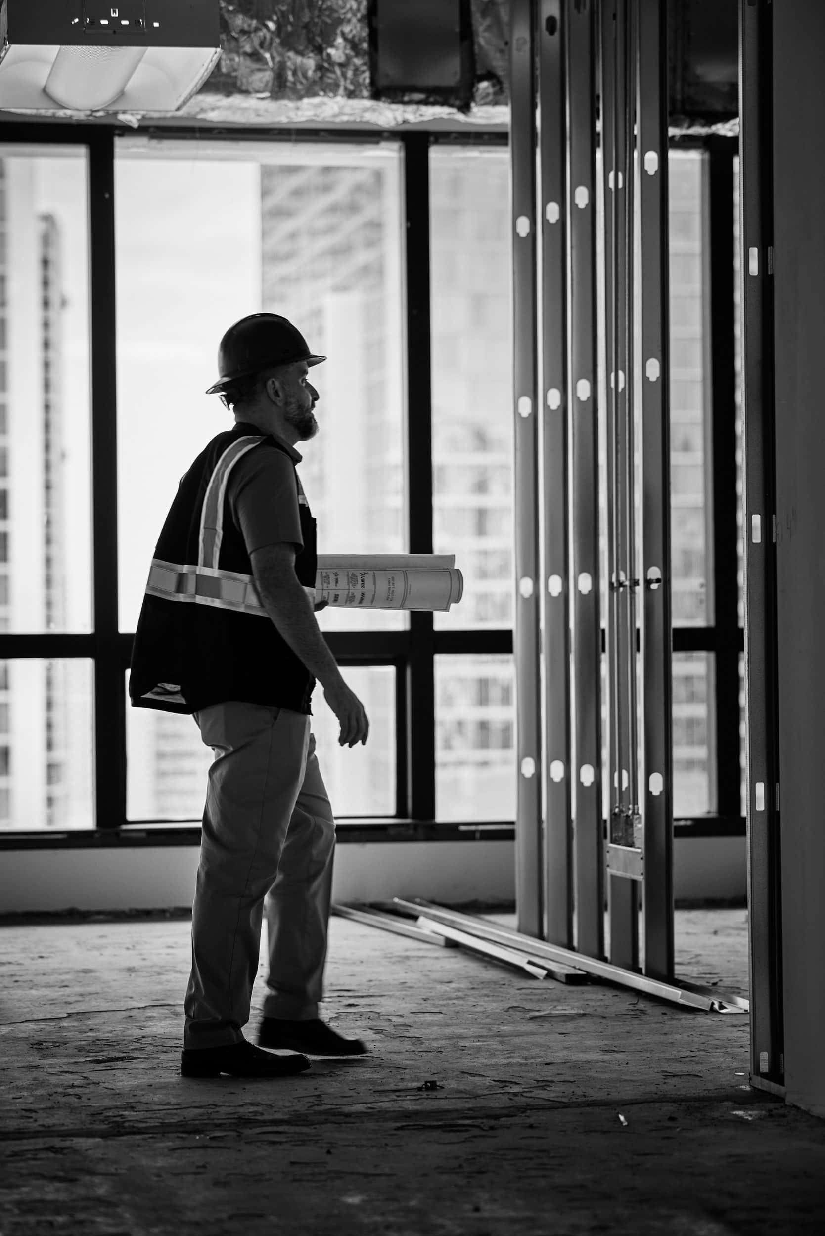 A project manager carrying rolled drawings surveys a floor under construction, seen in black and white