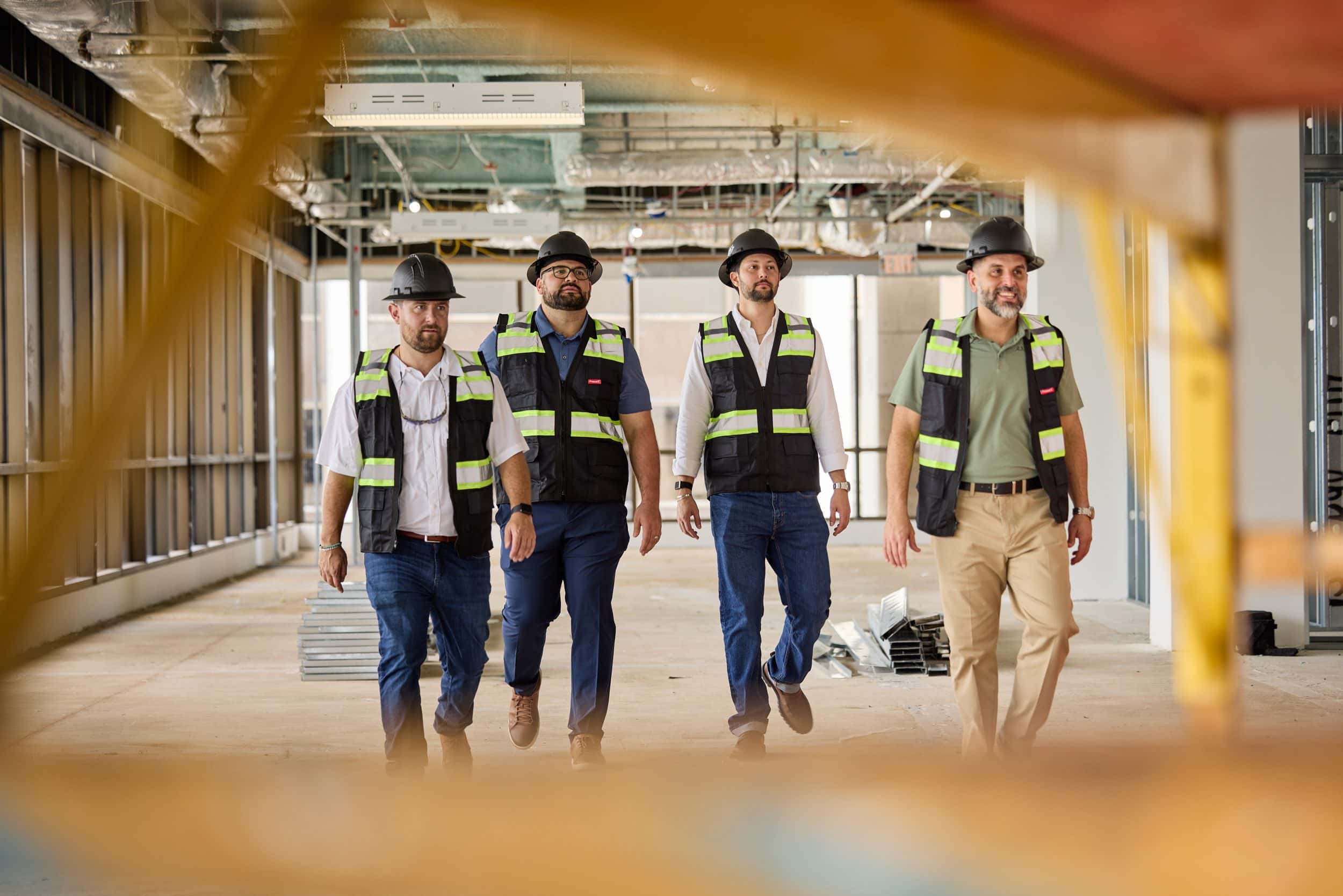 Four SPARC project managers in black hard hats and hi-vis vests walk toward the camera across a floor under construction